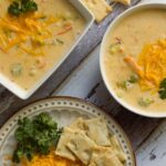 Two white bowls holding broccoli cheddar soup with a plate of toppings including crackers, parsley and cheddar cheese