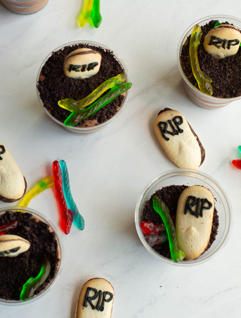 baileys chocolate mousse cups on a table with oreo dirt, gummy worms and tombstone cookies