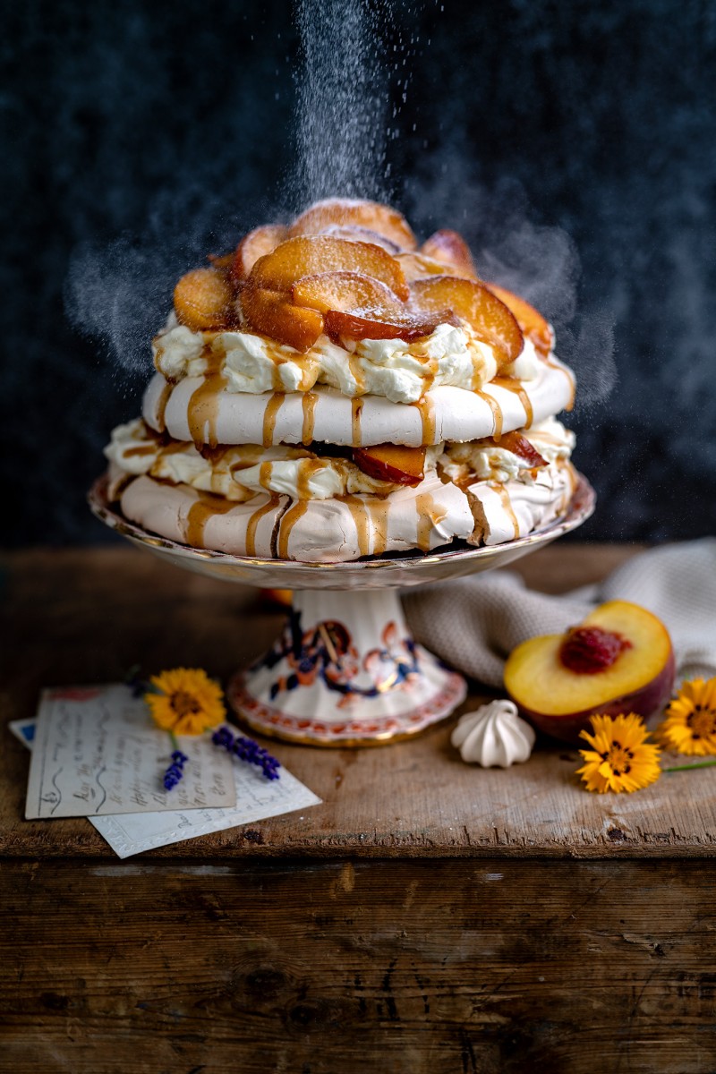 brown sugar pavlova on a stand with a dusting of powdered sugar against a dark backdrop