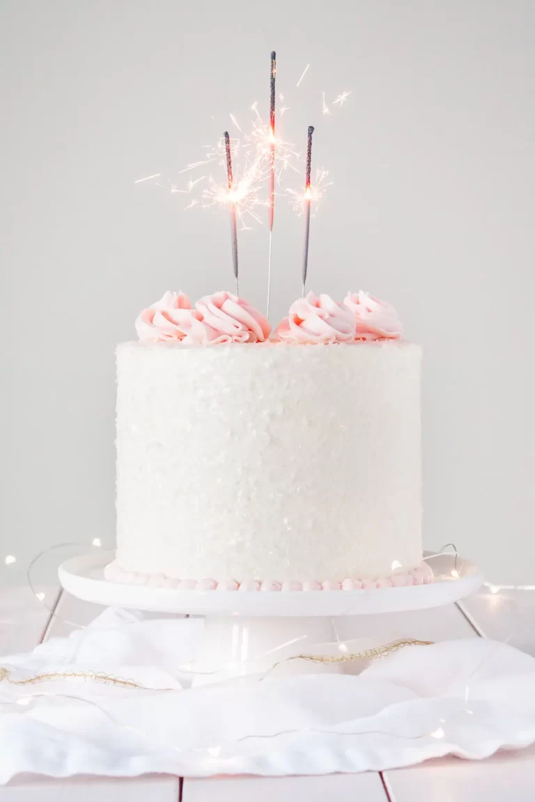 white cake with pink frosting on a white cake stand and pink sparklers on top