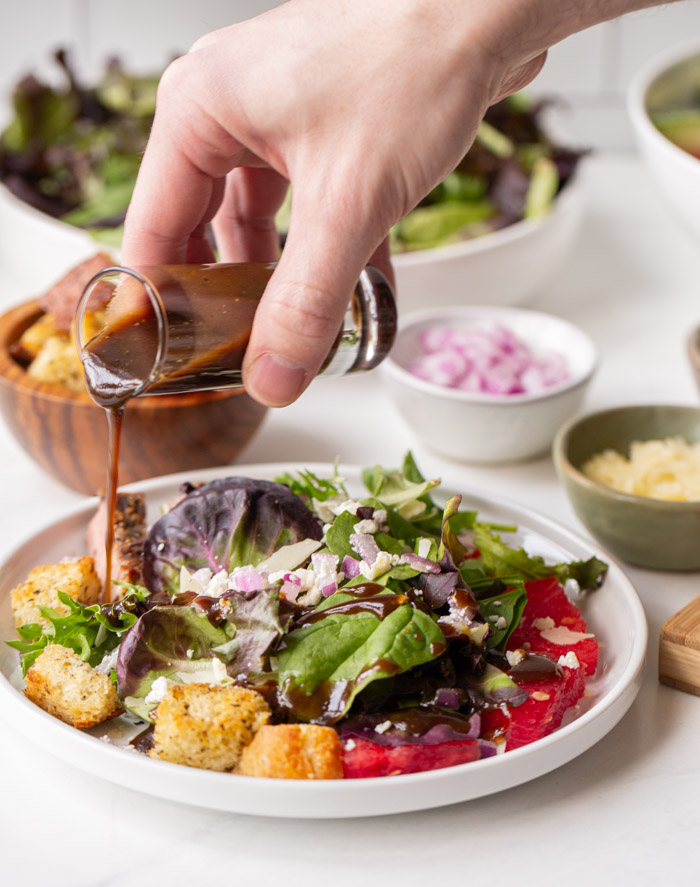 a salad containing steak, watermelon, red onion and feta with a hand pouring balsamic vinaigrette