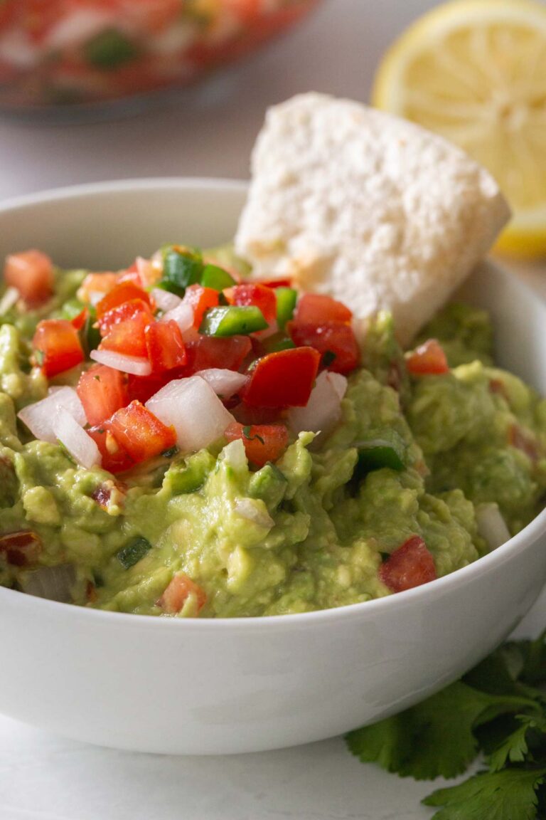 white bowl with guacamole and pico de gallo with a tortilla chip sticking out and a sliced lemon in the background