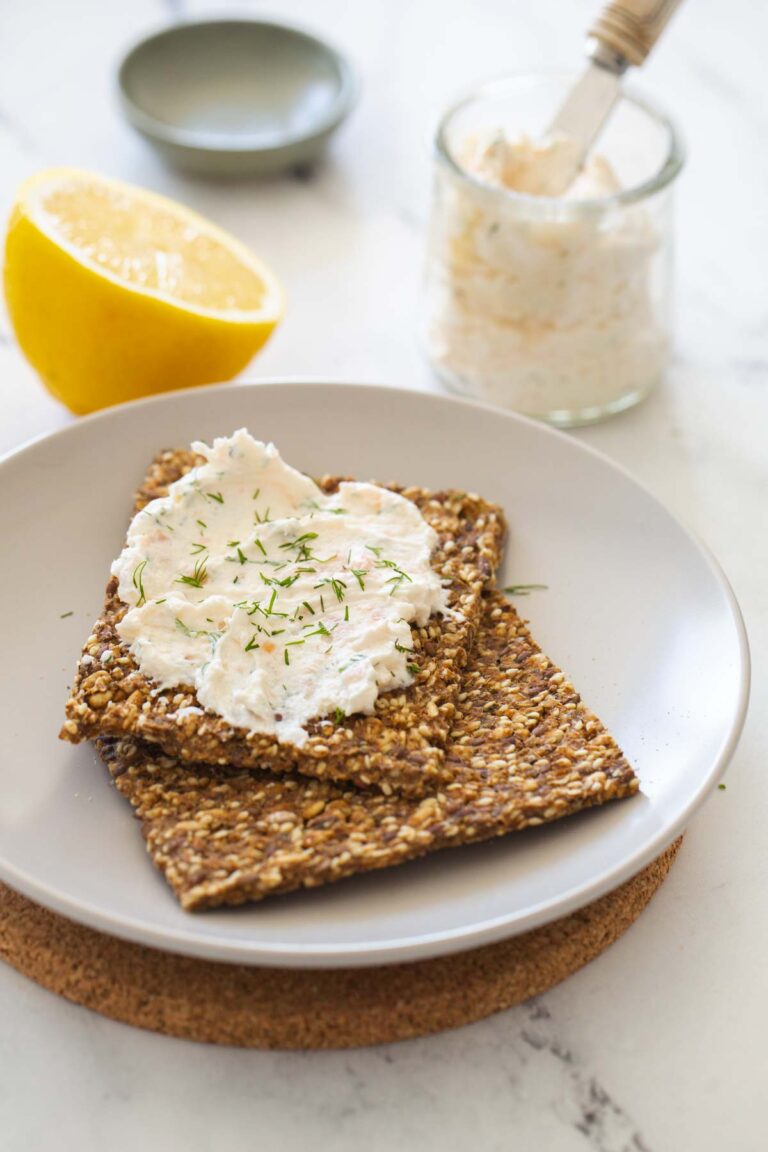 salmon mousse on seeded crackers topped with fresh dill on a grey plate on a marble background with a lemon and clear jar and small pinch bowl